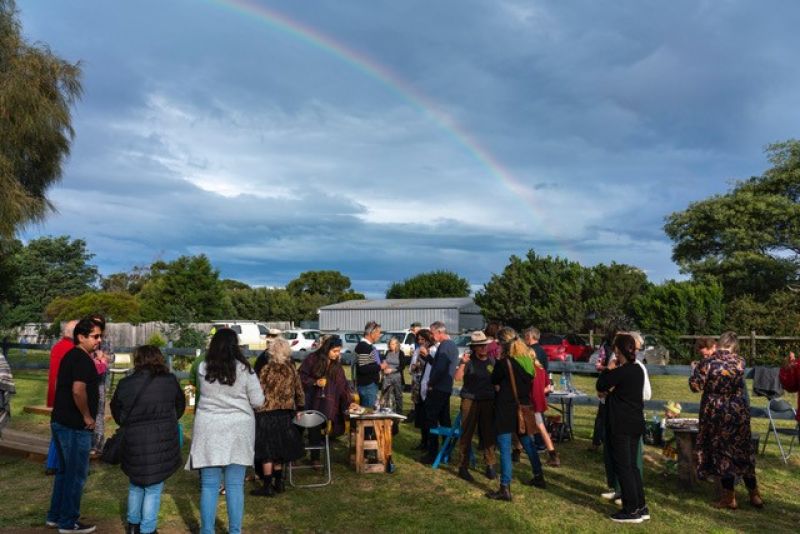 Neighbours gather to launch a new arts space on a sunny day in Marion Bay; a rainbow can be seen overhead.