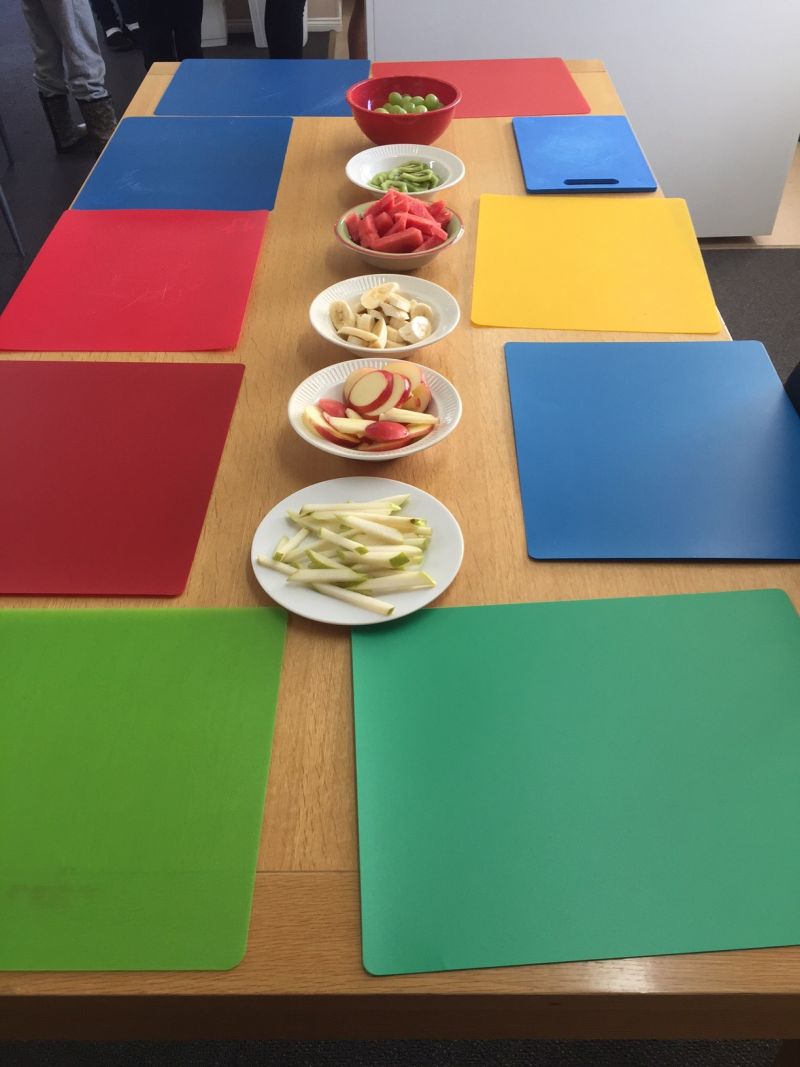 Table with brightly coloured placemats and healthy fruit and bowls of healthy fruit and vegetable in the centre of table.  Food Redi children’s program designed cooking games around ‘Superfood heroes’, covering a different hero each week.
